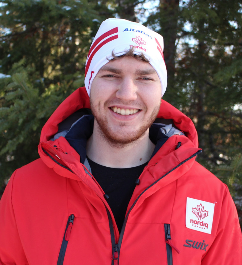 Jesse Bachinsky pose pour une photo, tout sourire. Il porte une veste de ski rouge vif, un t-shirt noir et une tuque blanche.