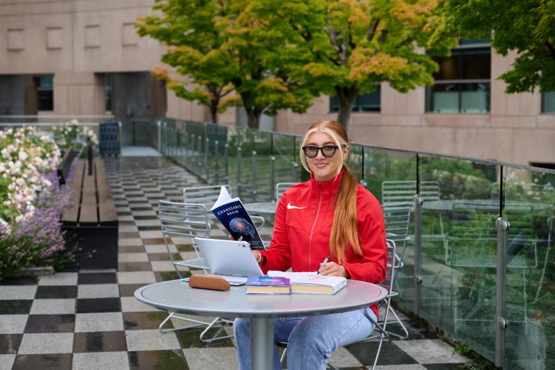 Ashlyn est assise à une table à l'extérieur d'un bâtiment scolaire, avec un ordinateur portable, des livres et des cahiers. Elle porte une veste rouge et des lunettes de soleil, et sourit à la caméra.