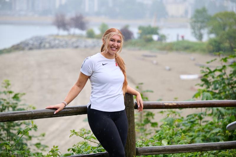 Ashlyn se tient debout à l'extérieur, posant devant une clôture en rondins. Elle sourit à la caméra et pose son bras gauche sur la clôture. Elle porte un haut de sport blanc et un pantalon noir.