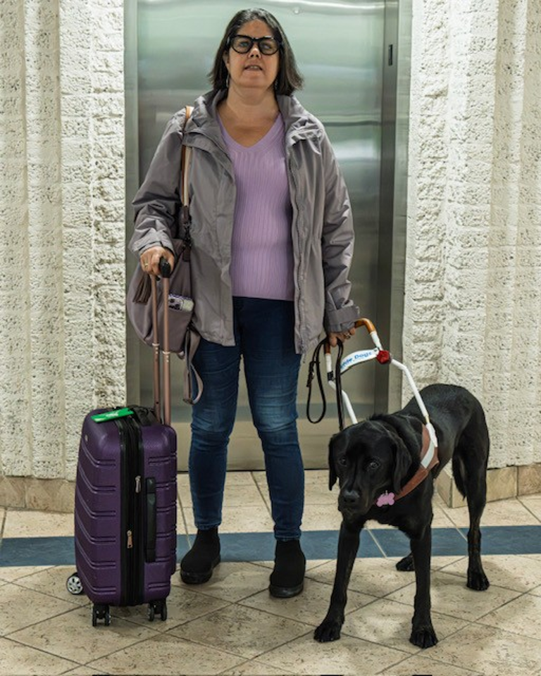 Karoline se tient à côté de son chien-guide, un labrador noir nommé Guardian, devant un ascenseur. Guardian porte son harnais ; Karoline tient la poignée du harnais dans sa main gauche et la poignée d’une valise dans sa main droite.
