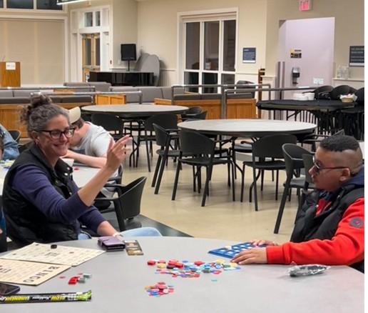 Sarah (center, wearing glasses and a dark vest) smiles warmly while assisting a guest during Bingo Night at CNIB Lake Joe. The two share a lighthearted moment as they play, surrounded by colorful game pieces scattered across the table.