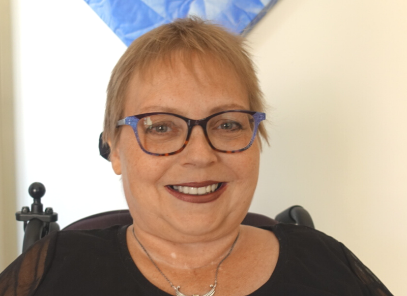 Un portrait de Lena souriant à l'appareil photo. Elle a les cheveux courts blond clair, porte des lunettes à monture bleue, un haut noir et un collier en argent, et est assise à l'intérieur