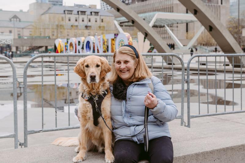  Lindsay est assise à côté de son chien-guide, Charles, un golden retriever, sur la place Nathan Phillips à Toronto. Charles porte son harnais et Lindsay sourit à la caméra, un bras passé autour de lui. 