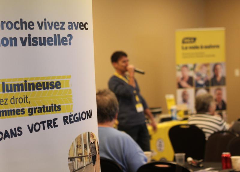 Une personne tient un micro et s'adresse à un public assis dans un Hub mobile d’INCA. Des bannières d'information aux couleurs de l'organisme sont visibles à l'arrière-plan.