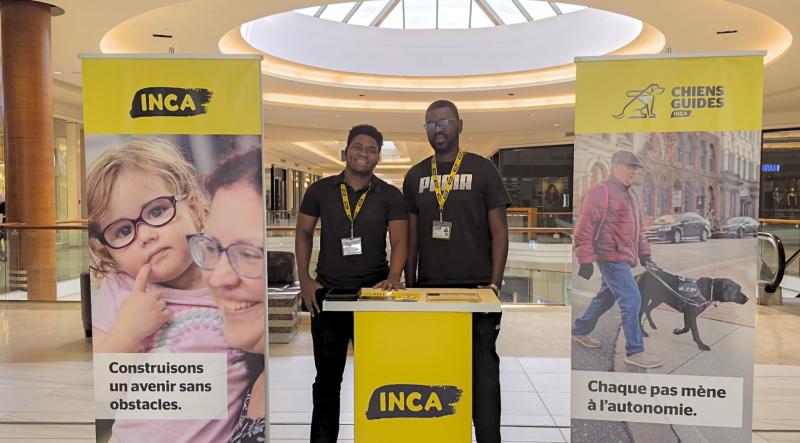 Deux solliciteurs portant des cordons d’identification d'INCA se tiennent près d’un kiosque d'INCA, à côté de bannières d'INCA et de l’association des Chiens-guides d'INCA, à l’intérieur d’un centre d’achats.