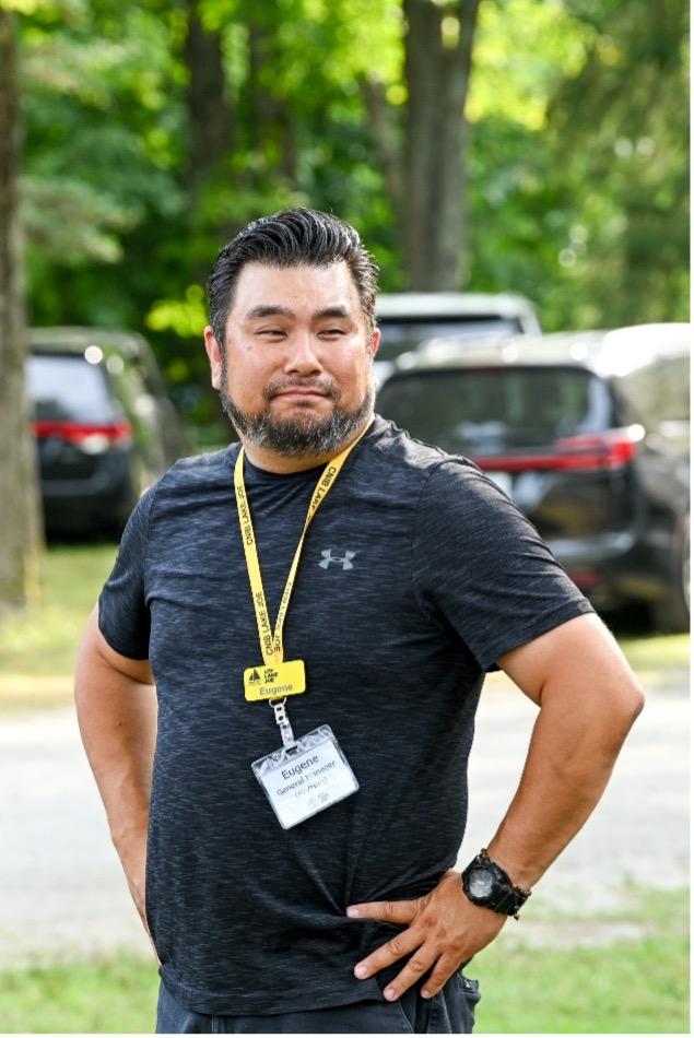 Eugene Chong se tient à l’extérieur, les mains sur les hanches, un léger sourire aux lèvres. Il a les cheveux courts, foncés et peignés vers l’arrière, ainsi qu’une barbe poivre et sel soigneusement taillée. Il porte un chandail de sport foncé, un cordon jaune du Camp Lake Joe d’INCA muni de badges nominatifs, ainsi qu’une montre-bracelet noire. Derrière lui, les arbres verts baignés de soleil et les véhicules stationnés apparaissent légèrement flous.