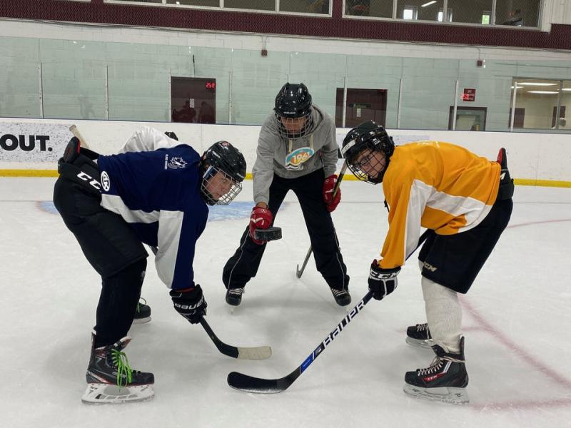 Deux joueurs de hockey aveugles tiennent leurs bâtons contre la glace et attendent que l'arbitre, qui se tient entre eux, lance la rondelle.