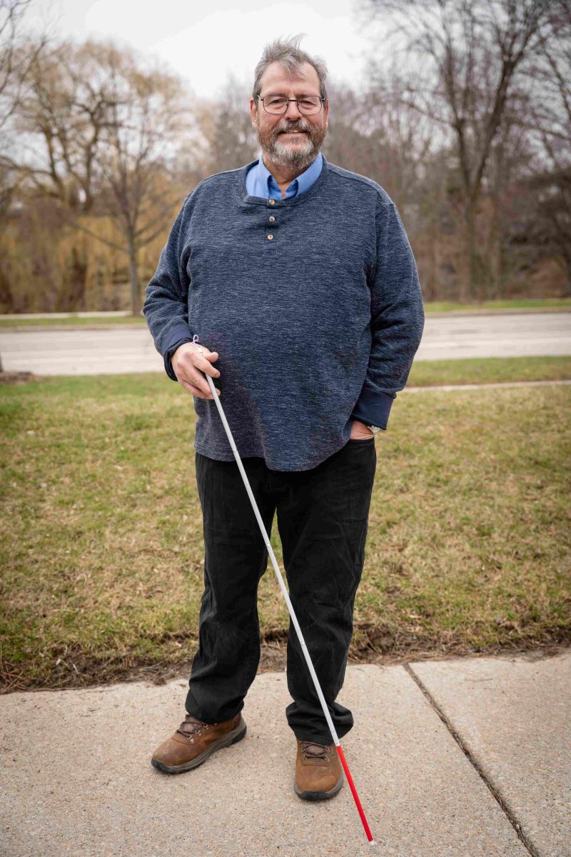 Un homme d'âge mûr, aux cheveux gris, portant des lunettes et une barbe, se tient debout en souriant sur un trottoir à l'extérieur. Il porte un pull Henley bleu marine par-dessus une chemise à col bleu clair, ainsi qu'un pantalon noir et des bottes marron. Il tient dans sa main droite une canne blanche à embout rouge.