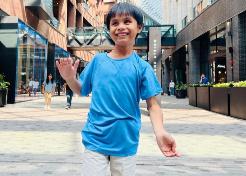 Un enfant souriant, vêtu d'une chemise bleue et d'un short clair, se tient dans une zone commerciale en plein air et fait signe de la main.