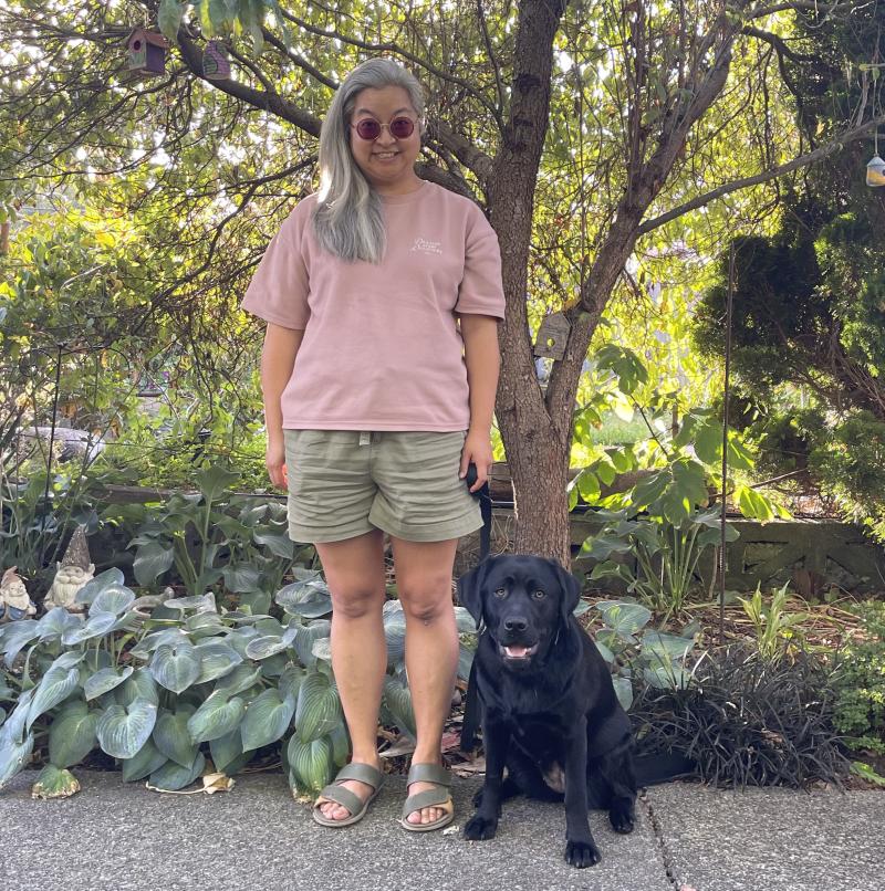 Theresa, vêtue d'une chemise rose et d'un short vert clair, se tient à côté d'un labrador noir, Summer, qui est assis. Theresa et Summer se trouvent sur un chemin devant un jardin avec des plantes vertes feuillues.