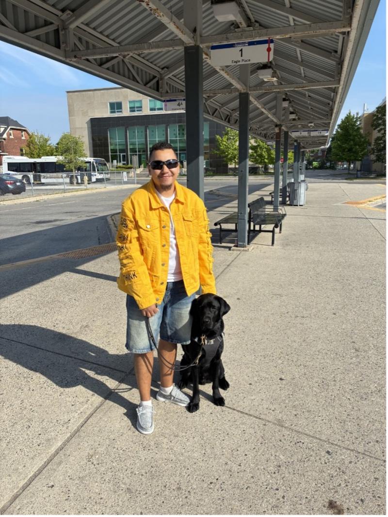 Tye se tient aux côtés de Zed, son chien-guide labrador noir, dans une station de transport en commun en plein air. Tye tient le harnais et la laisse de Zed.