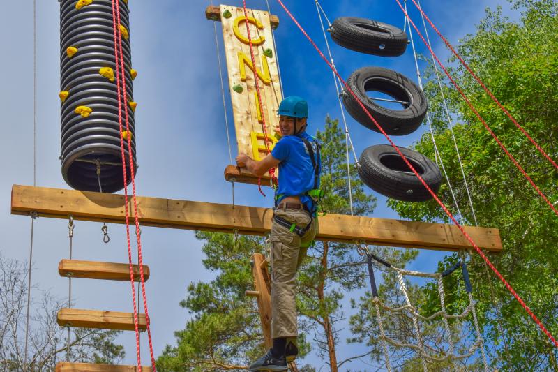 Une personne équipée d'un casque et d'un harnais d'escalade gravit l'aire de jeux verticale du CNIB Lake Joe, qui comprend des poutres en bois, des prises d'escalade, une échelle de corde et des pneus suspendus.