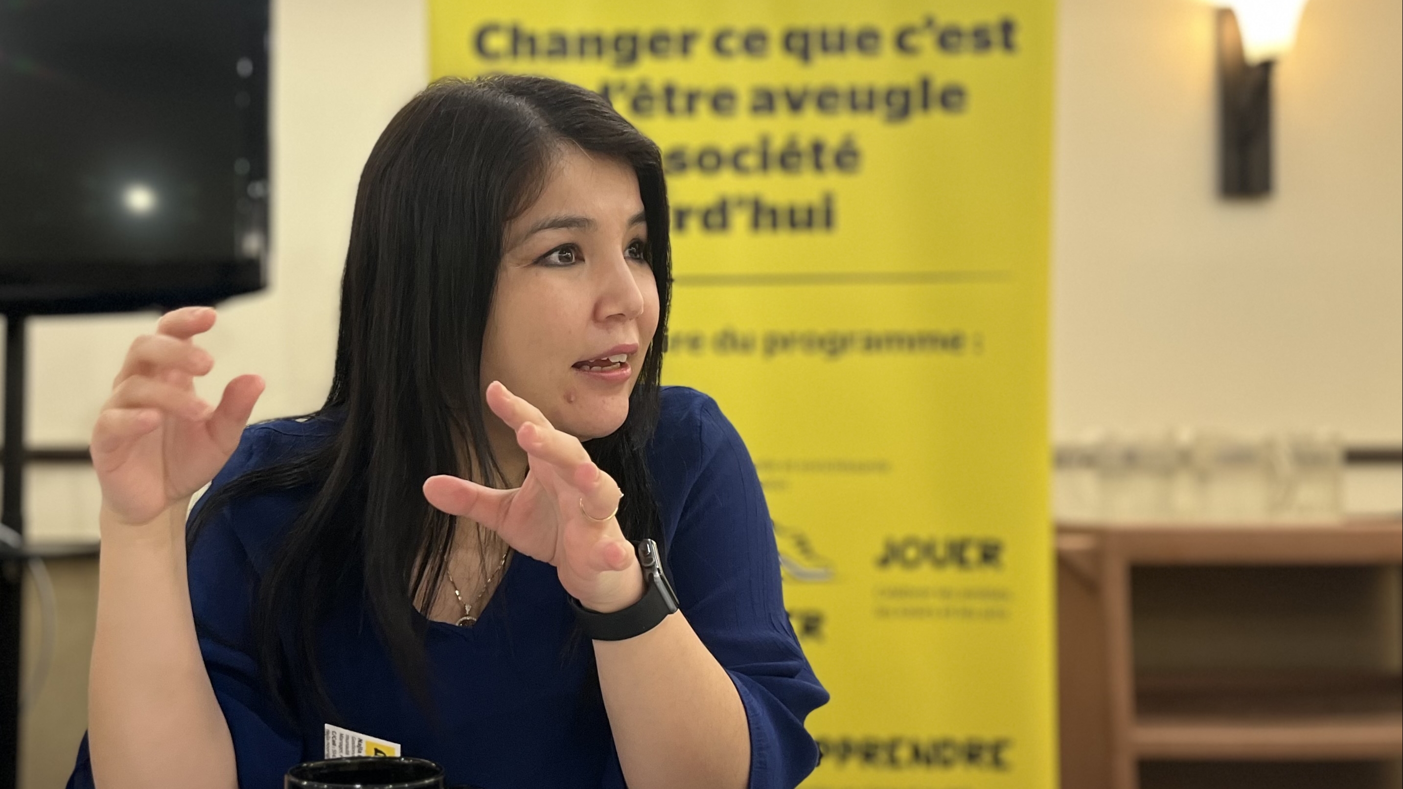 Najla parle avec ses mains, assise,  à un table, devant la bannière Changer ce que signifie être aveugle d'INCA.