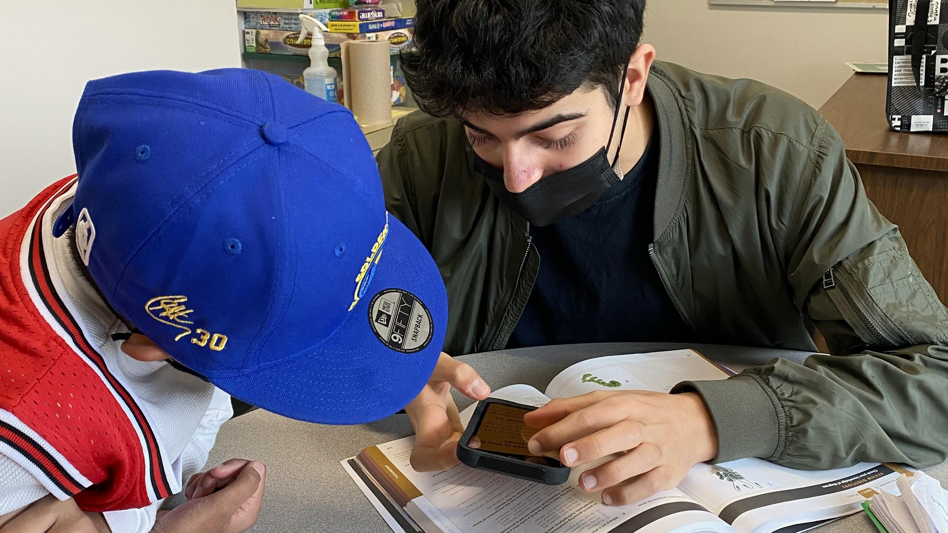 Dans une salle de classe, Aidan s’assoit avec un jeune élève et lui montre comment utiliser les fonctions d’agrandissement de son iPhone. Aidan tient son iPhone au-dessus d’un manuel scolaire. L’élève se penche attentivement pour assister à la démonstration.