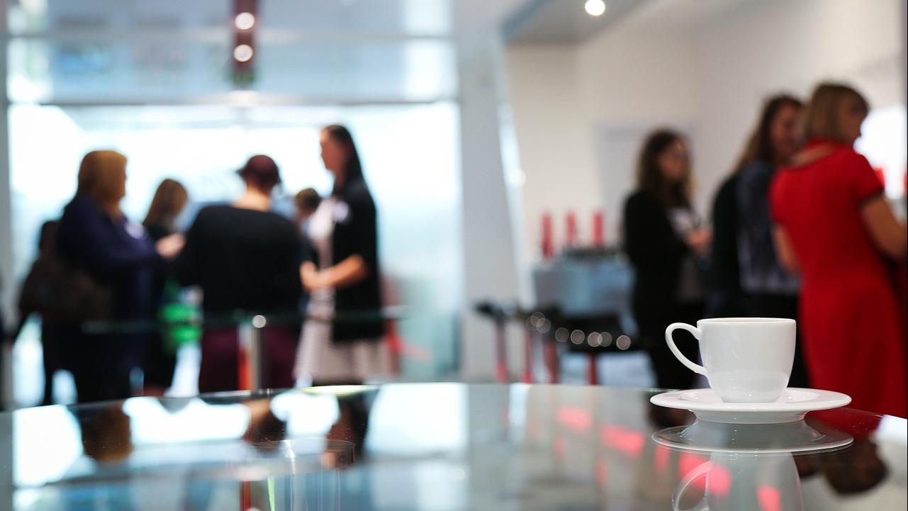 avant-plan d'une tasse de café, avec personnes qui discutent en arrière-plan