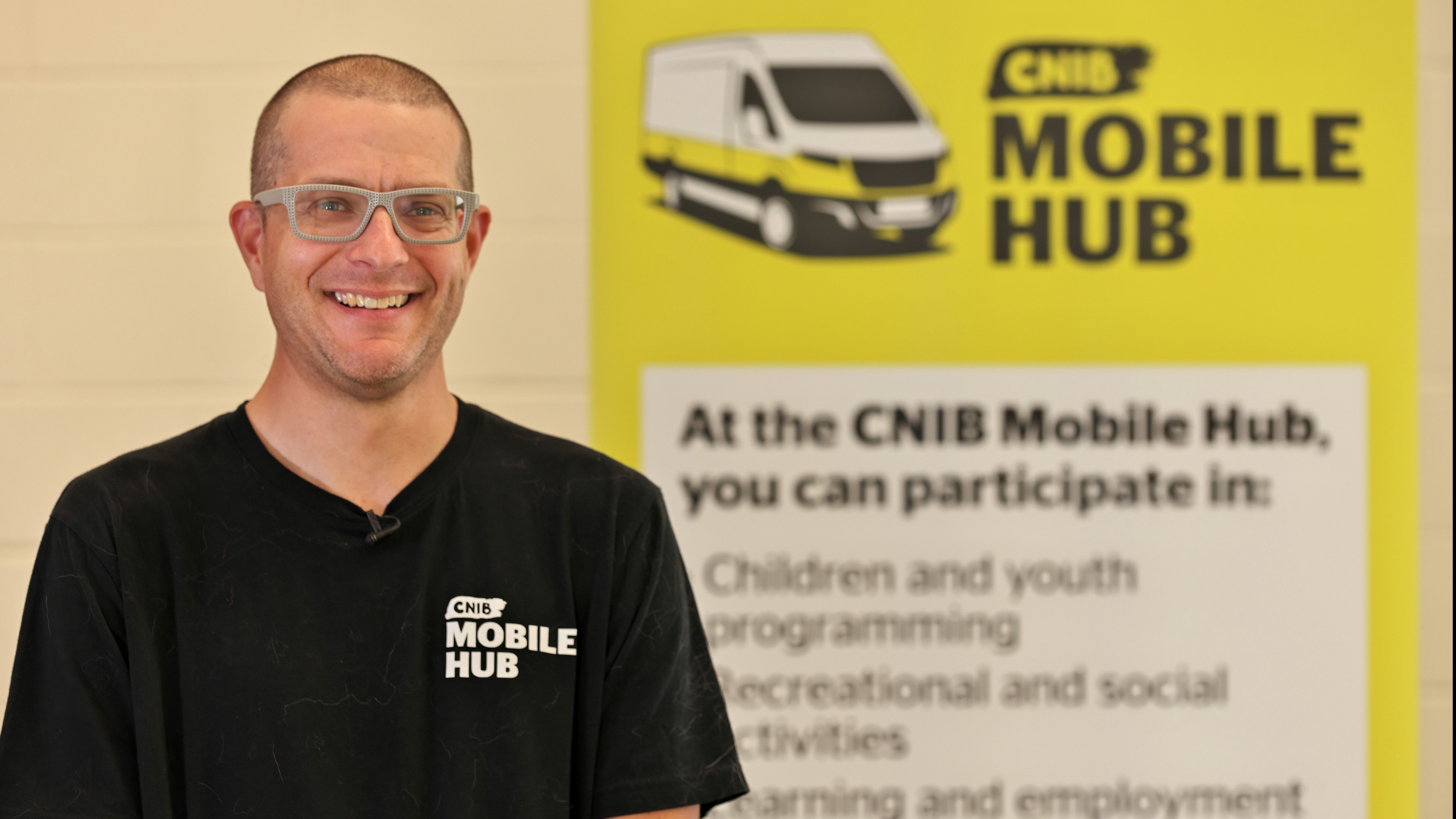 Plan moyen d'un homme à la peau claire et au crâne rasé, portant des lunettes et une chemise noire doté du logo Hub mobile CNIB, souriant à la caméra. On aperçoit à l'arrière-plan une bannière jaune du Hub mobile d'INCA.