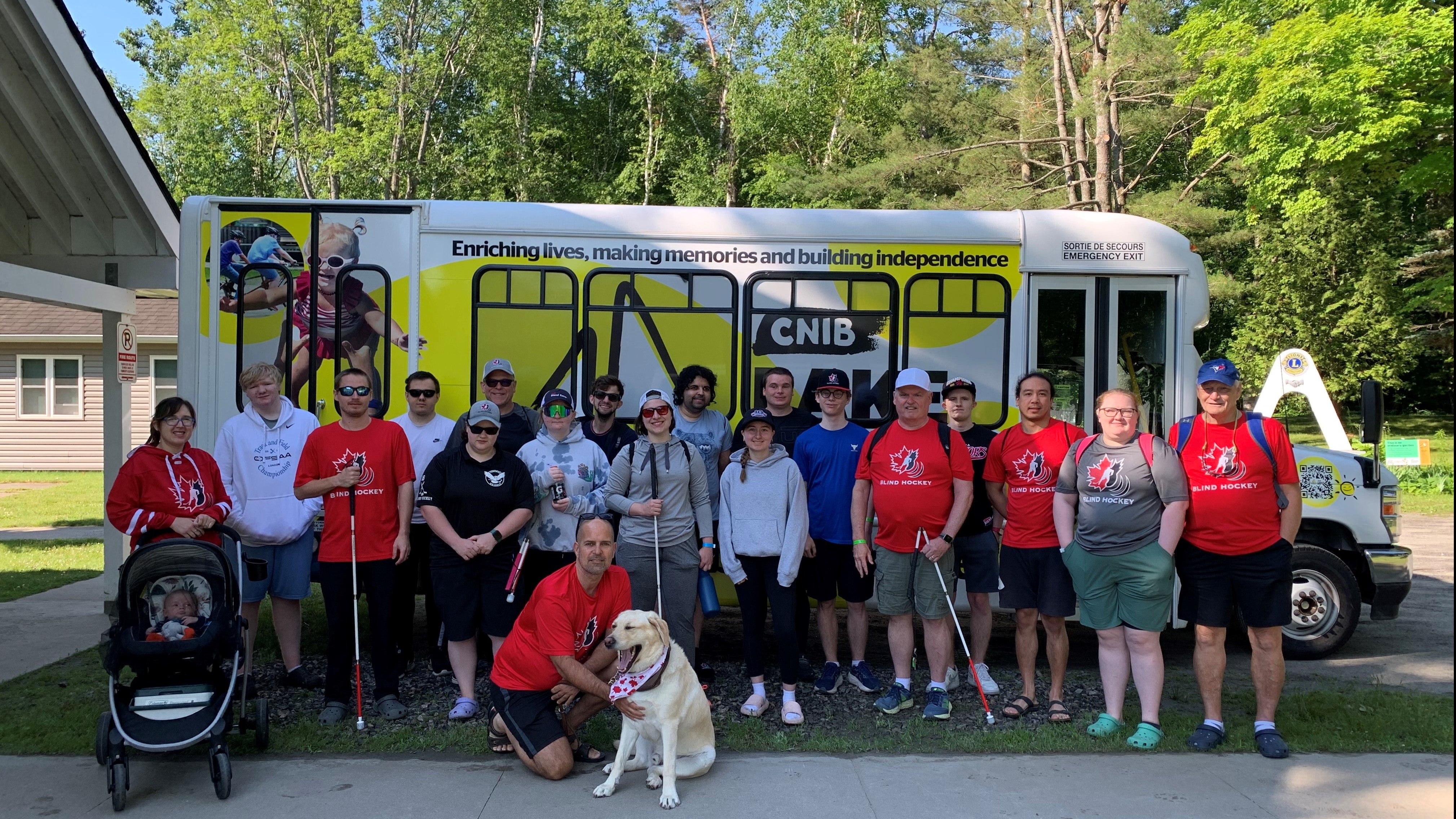 Environ 20 personnes se tiennent devant le nouveau minibus du Camp Lake Joe d'INCA. Plusieurs portent des t-shirts rouges du Hockey sonore Canada et tiennent des cannes blanches. Un chien-guide se trouve devant.