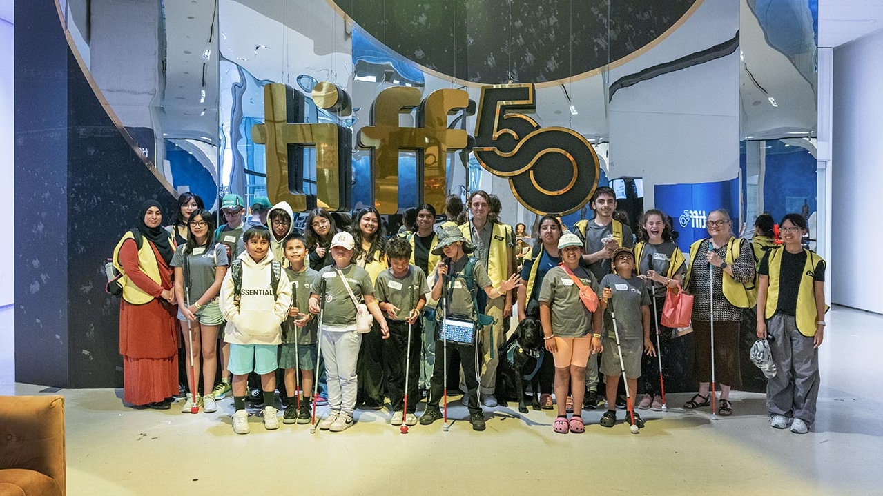 Dans le hall du TIFF Lightbox, un grand groupe de jeunes campeurs et de bénévoles d’INCA posent pour une photo de groupe devant une enseigne géante « TIFF 50 ».