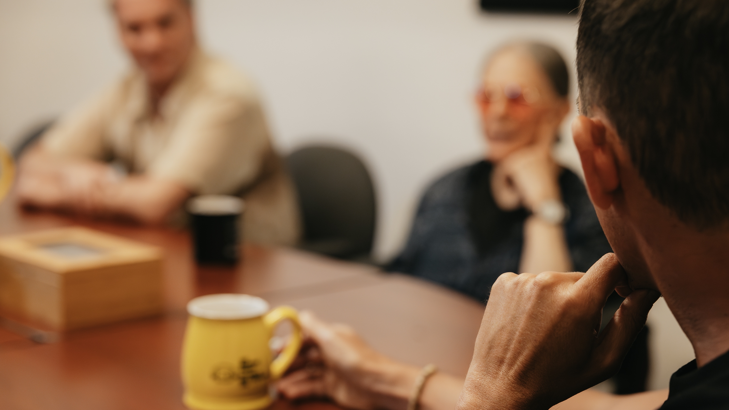 Trois personnes sont assises autour d'une table de conférence et discutent. L'attention se porte sur une personne au premier plan qui tient une tasse à café jaune. Deux autres personnes sont visibles à l'arrière-plan, légèrement floues, l'une portant des lunettes teintées de rouge et l'autre les bras croisés.
