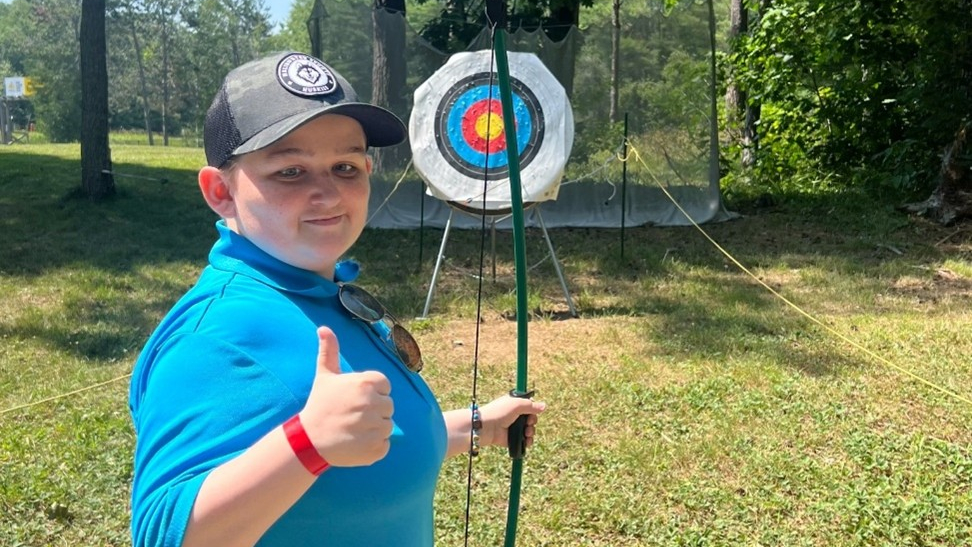 Un garçon tenant un arc et une flèche se tient devant une cible de tir à l'arc, levant le pouce en signe de victoire.