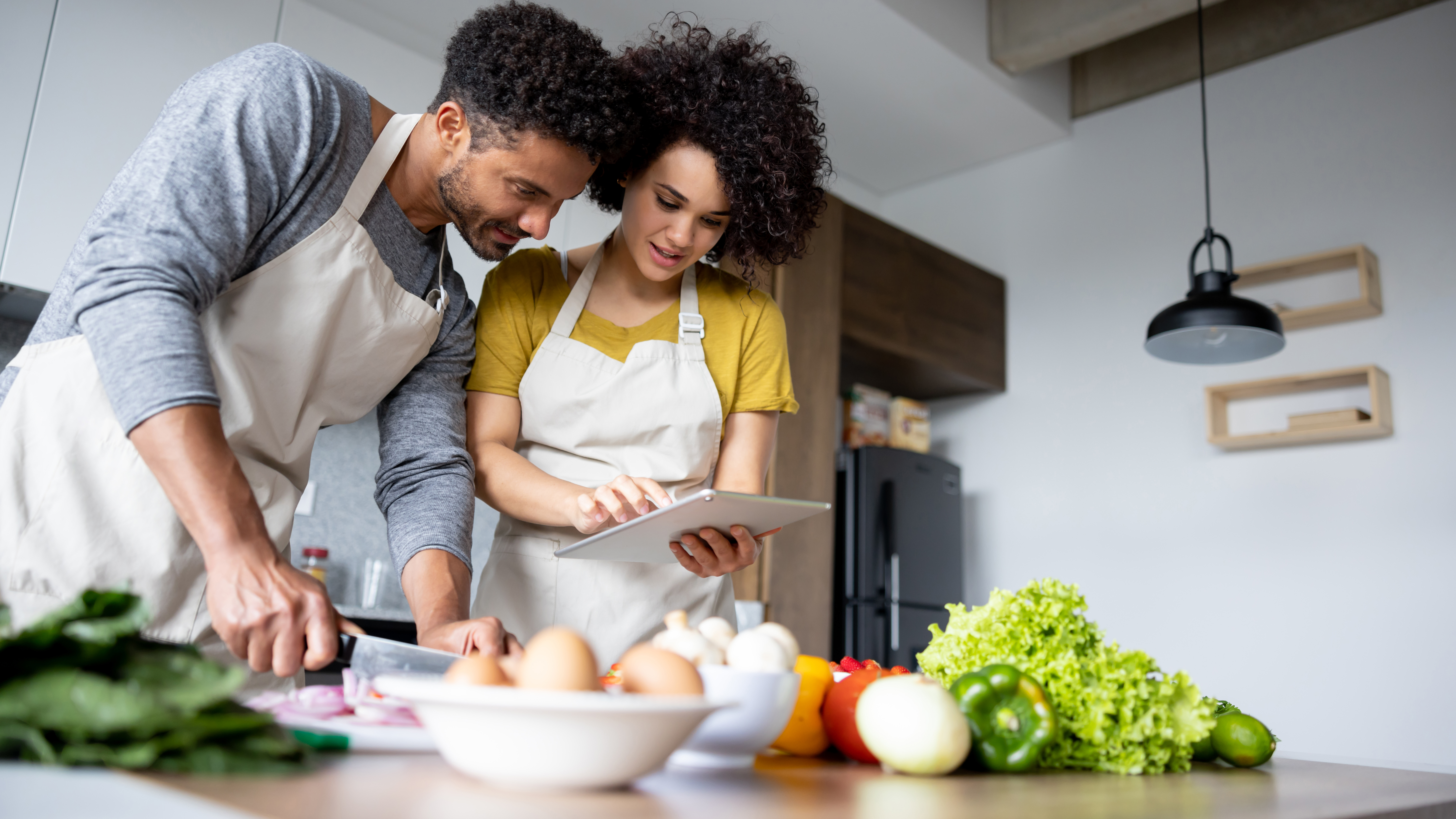Un jeune homme et une jeune femme portant un tablier, consultent une recette sur une tablette.  Le jeune homme coupe une partie des ingrédients qui sont devant eux.