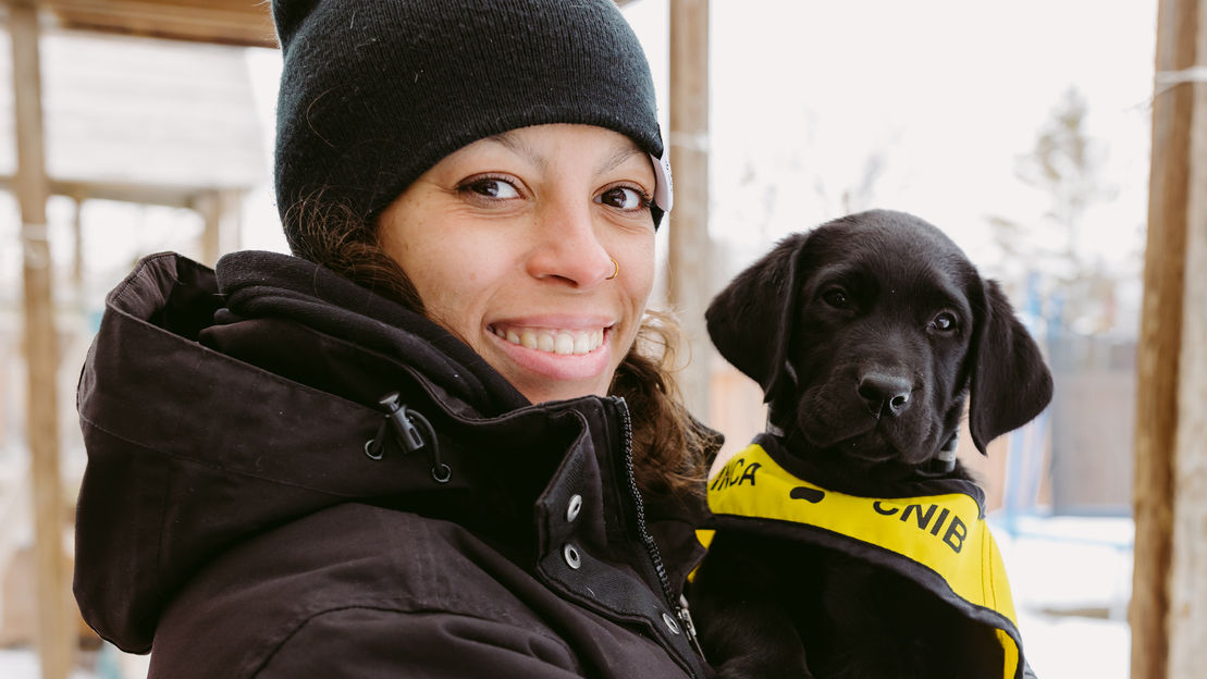 Une personne souriante, vêtue d'une veste d'hiver, tient dans ses bras un chiot noir portant un gilet du programme « Future Guide Dog » d'INCA.