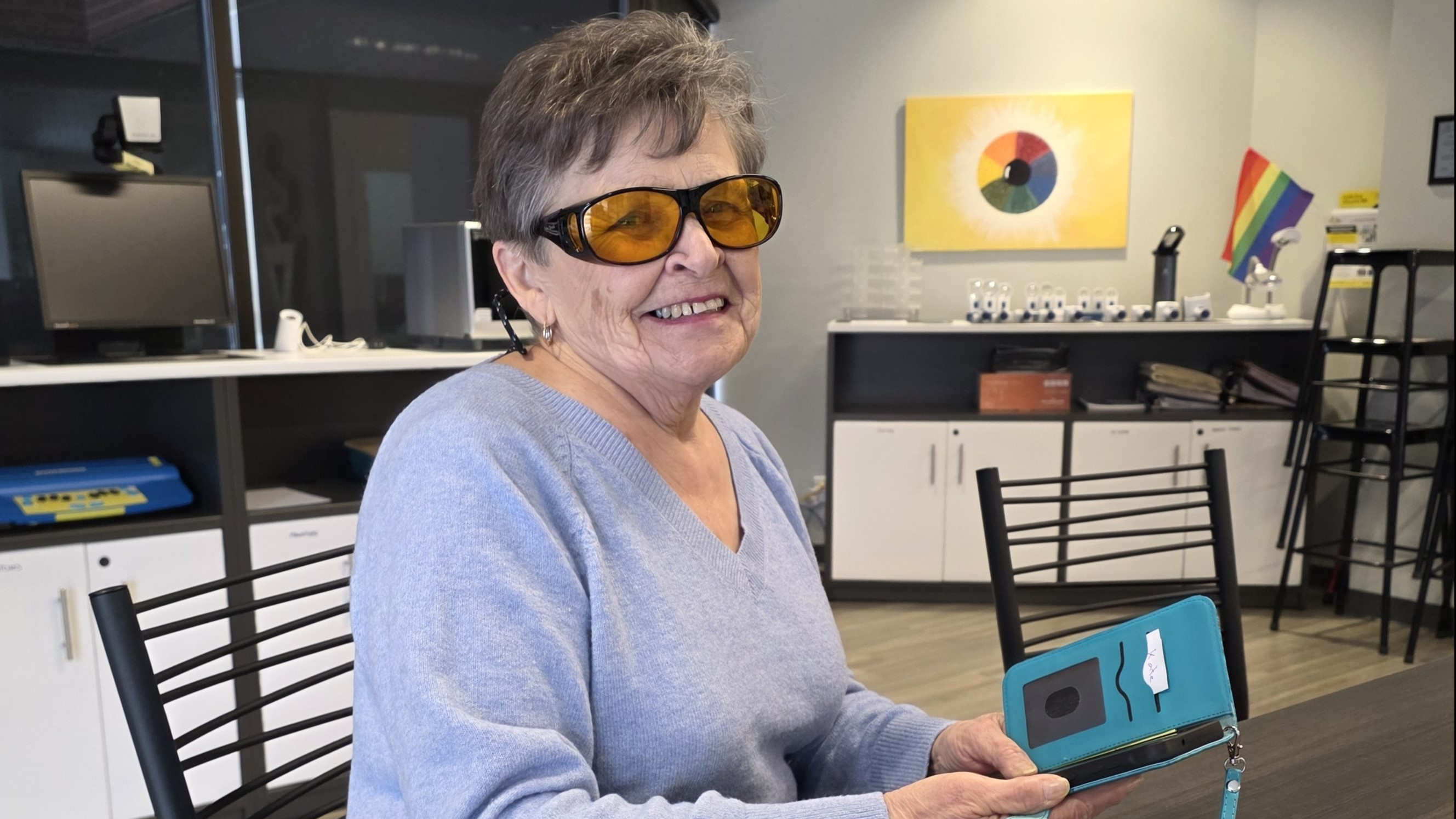 Une femme d'un certain âge portant des lunettes aux verres ambrés et tenant un téléphone intelligent adapté dans un étui bleu sarcelle.
