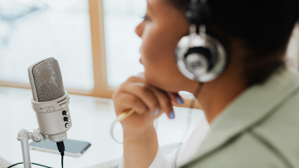Une femme portant un casque audio est assise à un bureau, à côté d'un micro sur pied. Elle est en train d'enregistrer un podcast.
