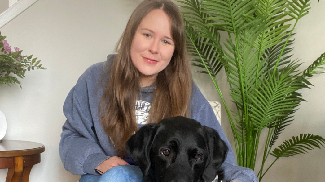 Taylor, une jeune femme aux longs cheveux bruns, est assise sur une chaise, le bras autour de son chien-guide Wallace, un labrador noir portant un harnais d'INCA, qui est assis à ses côtés.