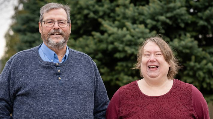 Un homme et une femme se tiennent côte à côte à l'extérieur, devant un grand conifère ; tous deux sourient et rient. L'homme, à gauche, a les cheveux gris, une barbe et porte des lunettes ; il est vêtu d'un pull Henley bleu marine par-dessus une chemise bleu clair. La femme, à droite, a les cheveux châtain clair et porte un haut texturé bordeaux foncé. L'image se présente sous la forme d'une grande bannière.