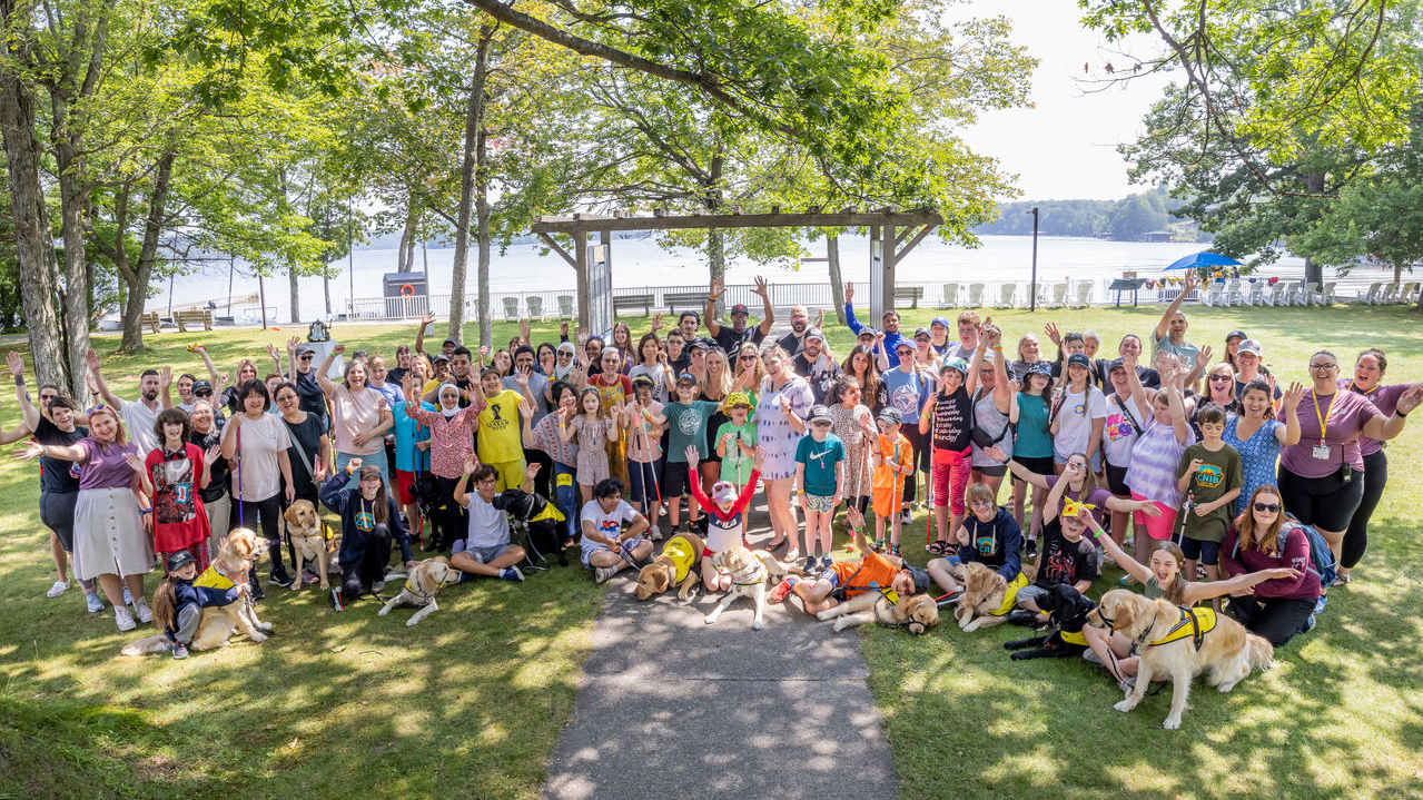 Au Camp Lake Joe, un large groupe de personnes de tous âges, dont bon nombre sont accompagnées de leurs chiens compagnons ou tiennent une canne blanche, lèvent joyeusement les mains en l'air, le lac en toile de fond.