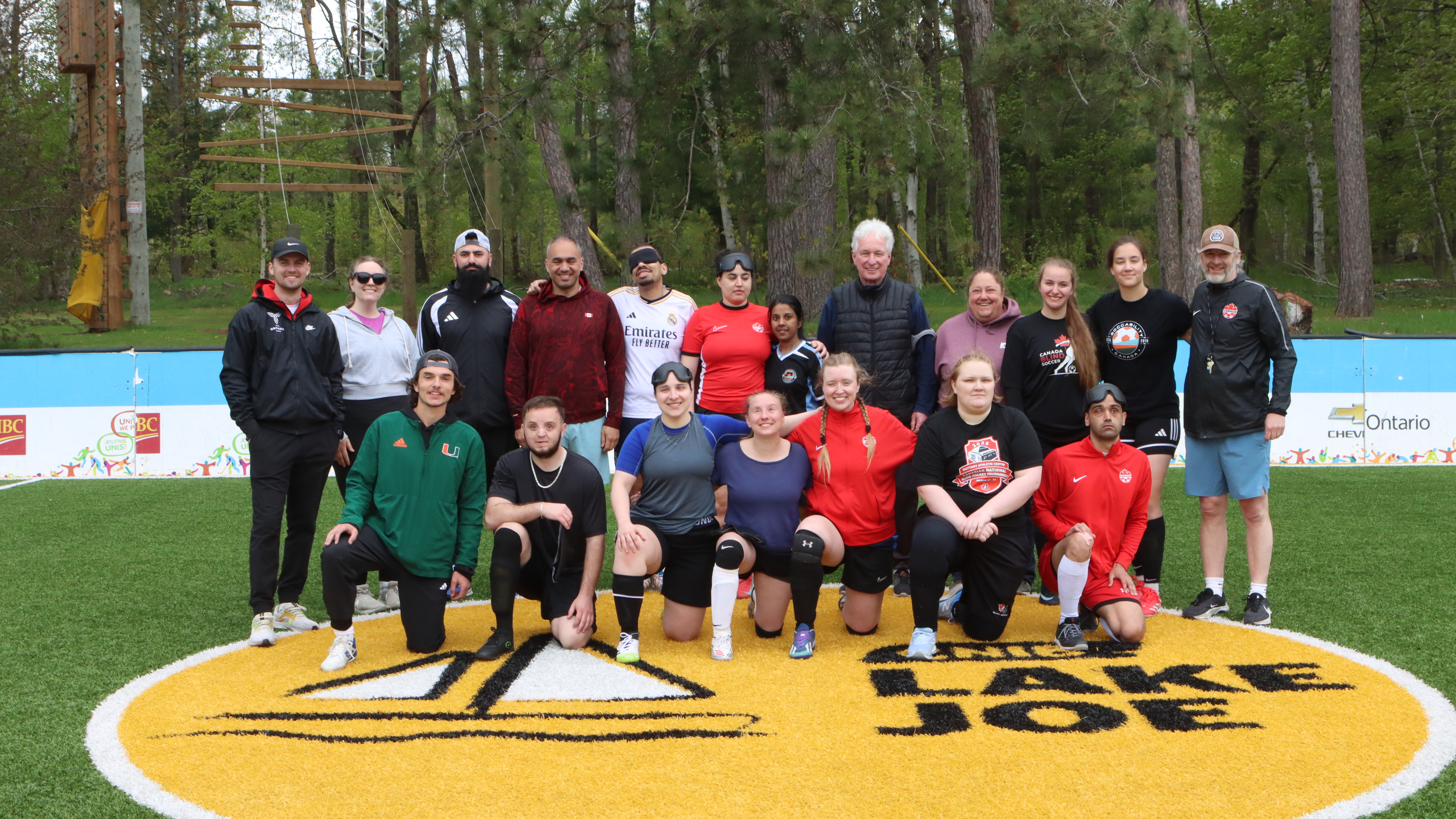 Un groupe de joueurs de soccer aveugles, de tailles, d'âges et de couleurs de peau variés en tenue de sport se tient derrière un ballon de cécifoot sur le terrain de soccer accessible du camp Lake Joe d’INCA.