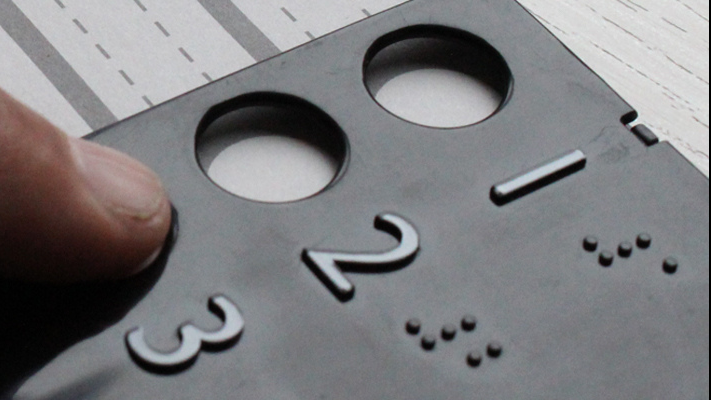 Gros plan sur une main qui explore au toucher un gabarit de vote tactile en relief, avec des chiffres 1, 2, 3 et leurs équivalents en braille, posé sur un bulletin de vote.