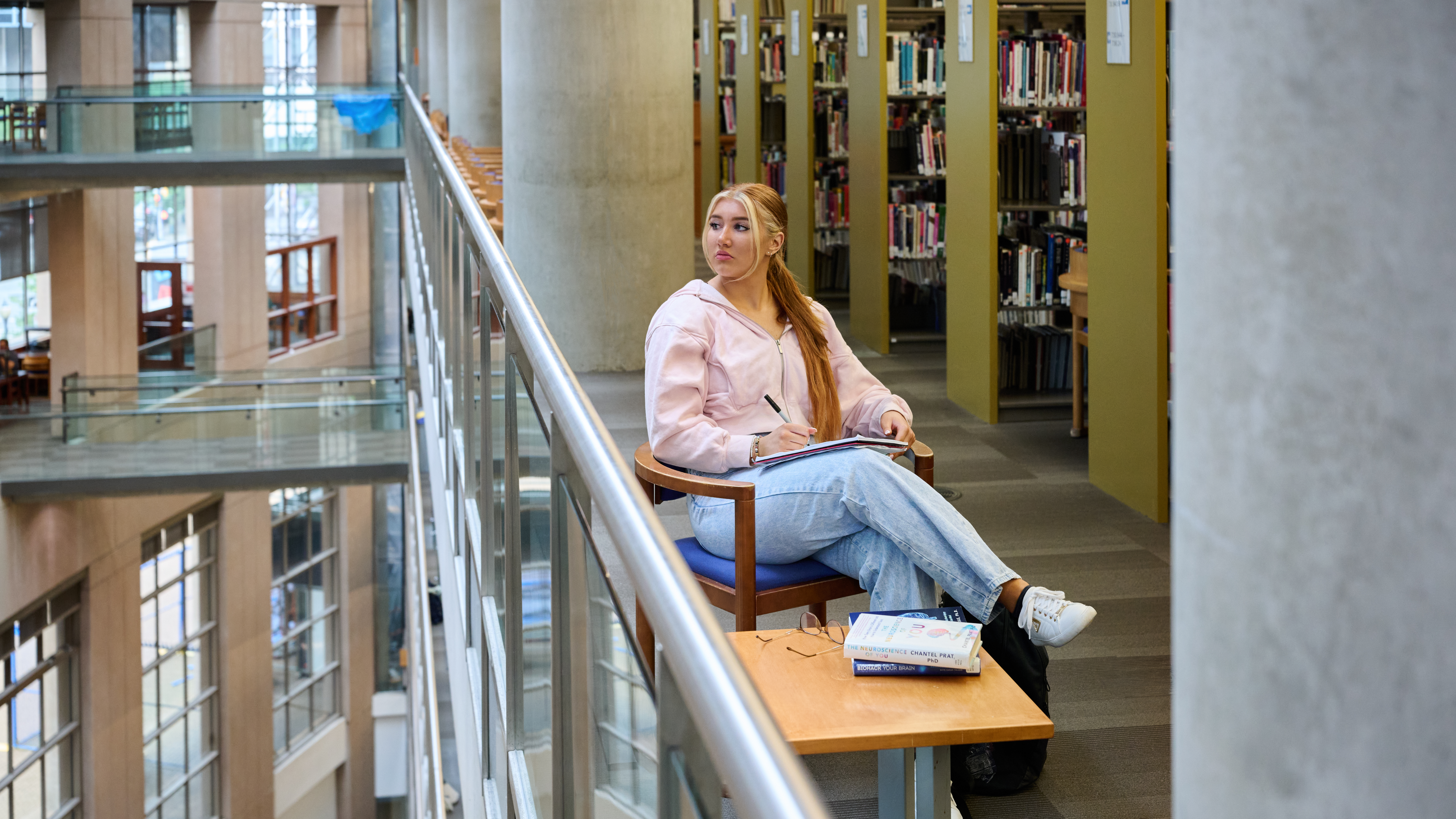 Ashlyn, vêtue d'un pull rose et d'un jean, est assise sur une chaise en bois près de la balustrade en verre d'une bibliothèque à plusieurs étages. Elle tient un cahier et un stylo sur ses genoux et regarde par-dessus le bord de la balustrade.