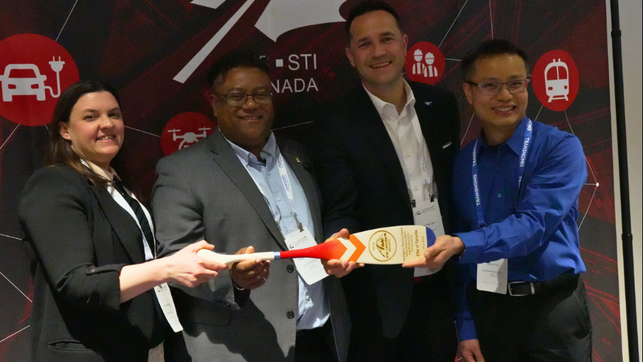 Quatre membres de l'équipe du projet pilote « Zones de construction de trottoirs accessibles aux personnes aveugles et à basse vision » posent ensemble lors d'un événement, souriants et tenant leur prix, une pagaie en bois sur laquelle sont gravées les informations relatives à cette distinction. Mary Ann, à l'extrême gauche, tient une extrémité de la pagaie tandis que les autres membres de l'équipe se rassemblent autour d'elle.