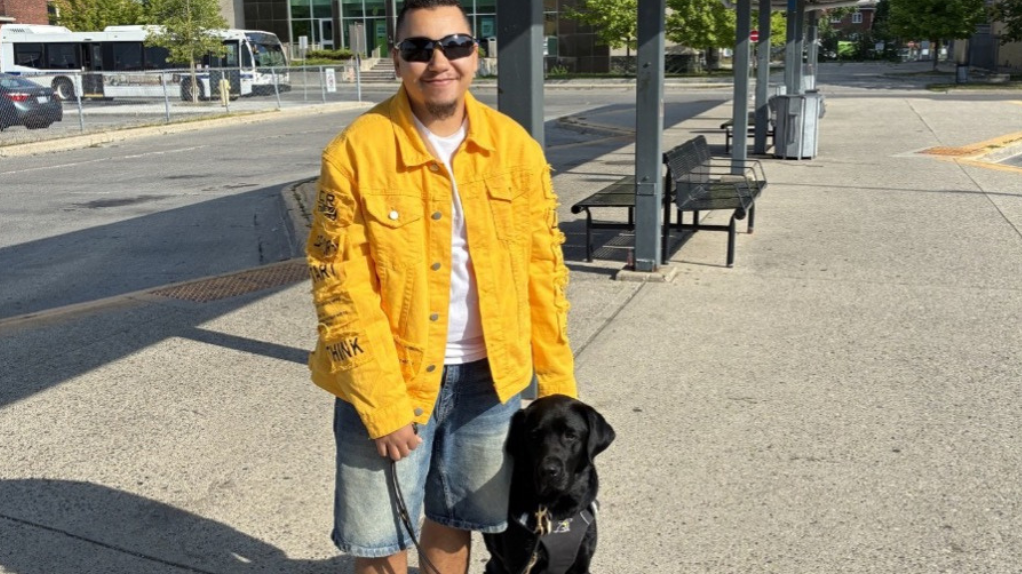 Tye se tient aux côtés de Zed, son chien-guide labrador noir, dans une station de transport en commun en plein air. Tye tient le harnais et la laisse de Zed.
