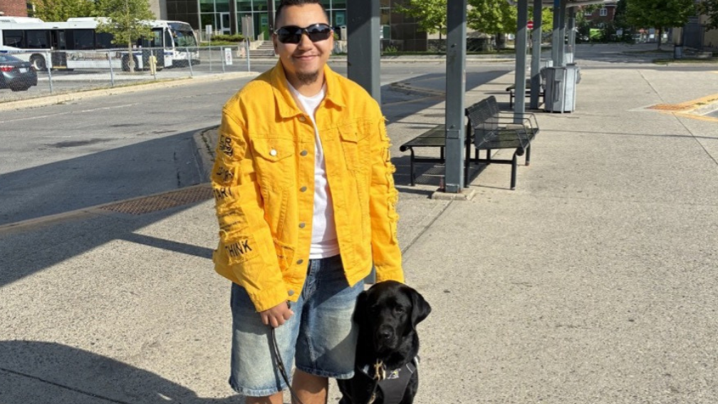 Tye se tient aux côtés de Zed, son chien-guide labrador noir, dans une station de transport en commun en plein air. Tye tient le harnais et la laisse de Zed.