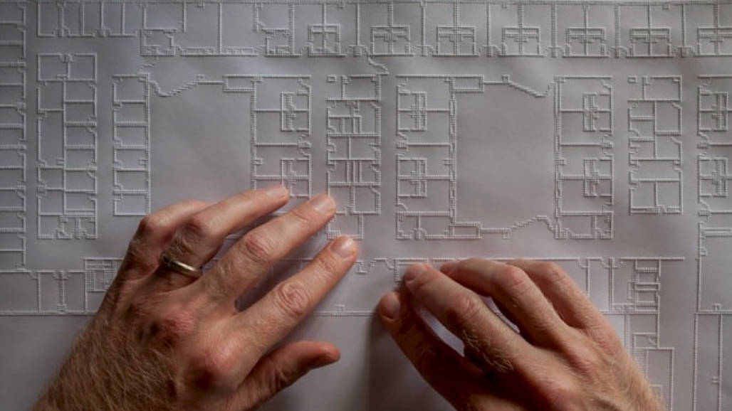 Deux mains parcourent un grand plan architectural tactile. Des lignes en relief délimitent les pièces, les couloirs, les murs et d'autres éléments de la disposition du bâtiment.