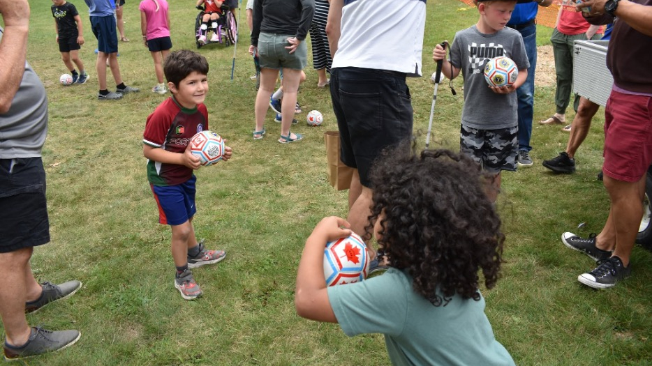 Une foule d’enfants et d’adultes se rassemble sur une pelouse pour une activité de football. Beaucoup d’entre eux tiennent de petits ballons de football blancs dans leurs mains ou les font rouler à leurs pieds.