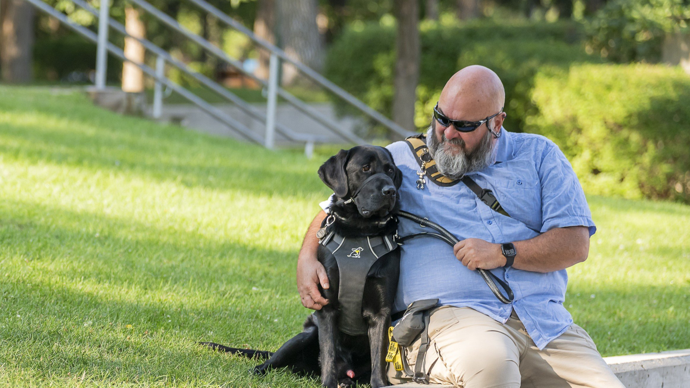 Jason est assis sur un rebord dans un parc, à côté de Midas, son chien-guide noir. Jason regarde Midas en souriant ; il porte un t-shirt bleu clair à col, un pantalon beige et des lunettes de soleil noires.