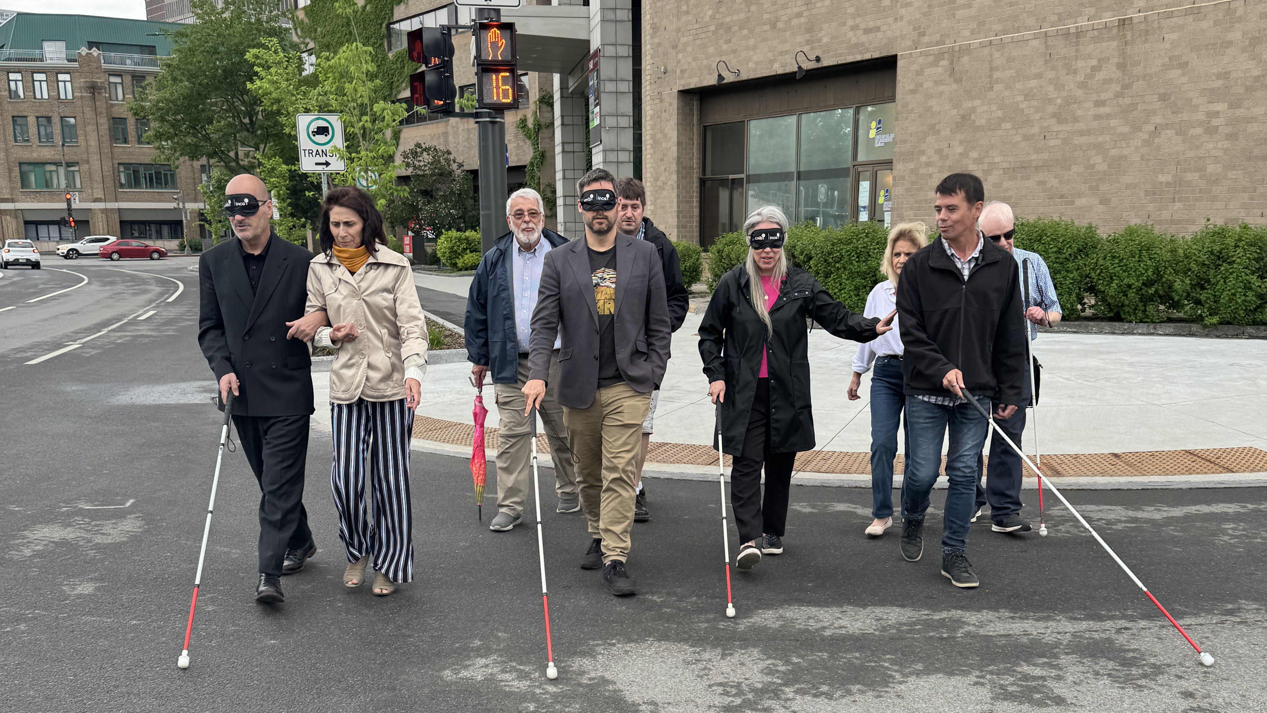 un groupe de personnes qui traversent la rue, y'en a qui ont les yeux bandés qui ont une canne et qui sont guidés par soit une personne ayant une limitation visuelle soit une personne voyante.