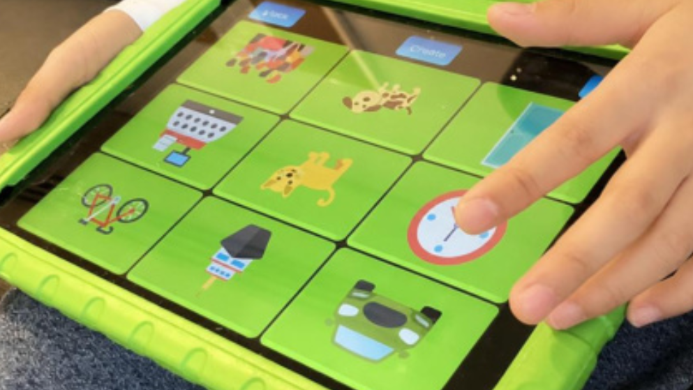 Les mains d’un enfant tiennent un iPad dans un étui de protection vert vif et tapotent une grille d’icônes colorées représentant notamment des animaux, des objets et une horloge à l’écran.