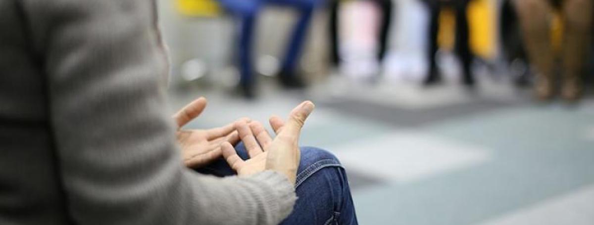 A person sits in a group circle. Their hands are clasped in their lap.