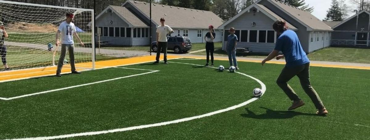 CNIB Lake Joe staff member kicking ball at goalie in the net with three people observing.