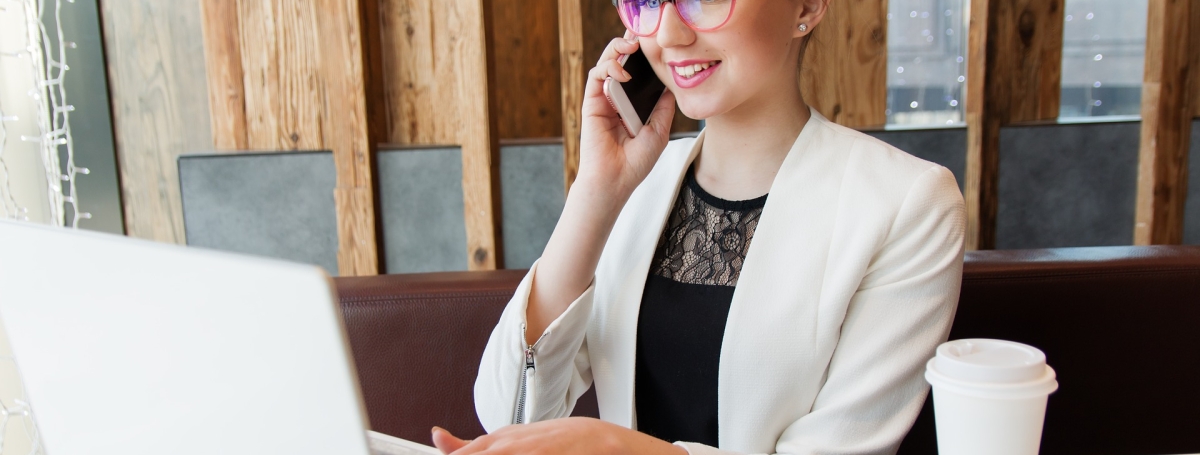 Une jeune femme  portant des lunettes parle au téléphone et est sur l'ordi. un café pour emporté est déposé sur la table. 