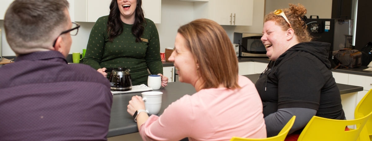 3 jeunes femmes et un jeune homme rient de bon coeur autour d'un bon café dans une cuisine.