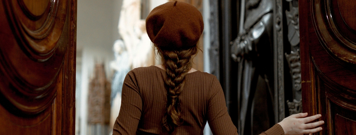 Une femme coiffée d’un béret brun entre dans une salle de musée ornée de sculptures en poussant une grande porte en bois.