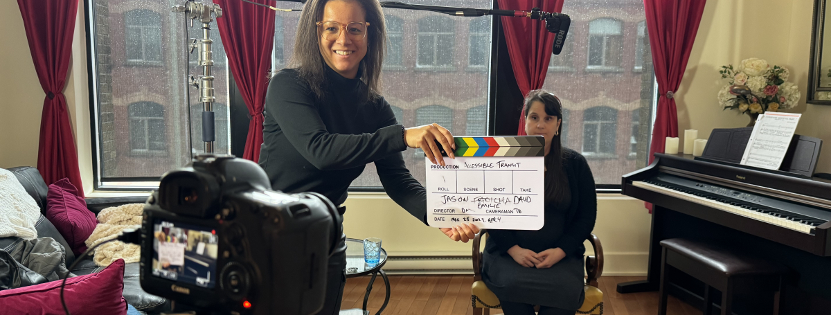 Une femme souriante qui tient le clap cinéma, on aperçoit une camera en avant plan et en arrière plan une femme assis sur une chaise