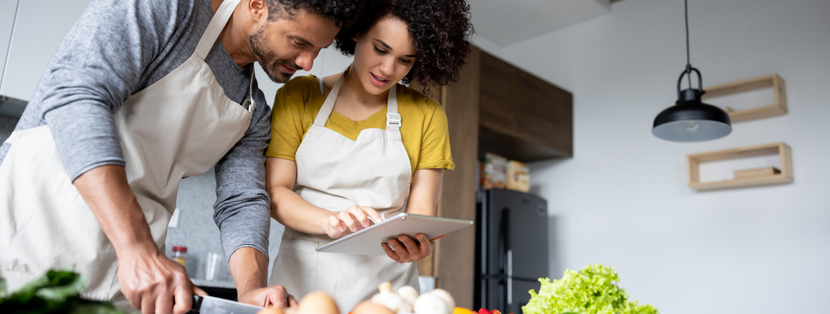 Un jeune homme et une jeune femme portant un tablier, consultent une recette sur une tablette.  Le jeune homme coupe une partie des ingrédients qui sont devant eux.