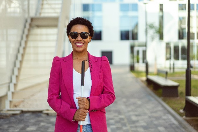 Une femme souriante portant un blazer et des lunettes de soleil tient une canne blanche.